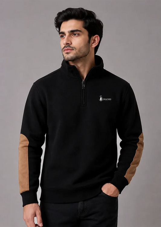 A man wearing a black quarter-zip pullover sweatshirt with tan color-blocked elbow patches and a small white embroidered logo on the left chest, posed against a solid gray studio background.