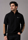 A man wearing a black quarter-zip pullover sweatshirt with tan color-blocked elbow patches and a small white embroidered logo on the left chest, posed against a solid gray studio background.