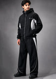 A man modeling a black hooded zip-up jacket and matching wide-leg track pants with white side panels and piping, photographed against a dark gray studio background.