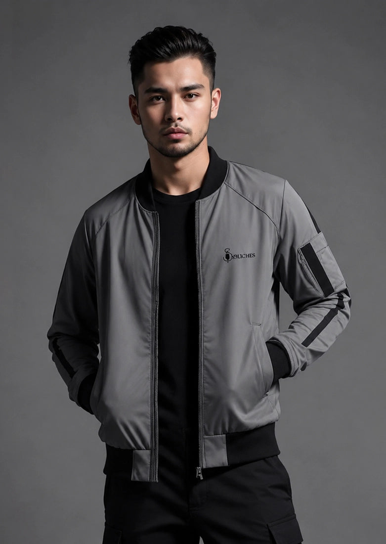 A man wearing a gray and black bomber jacket with a black crewneck shirt and black pants, standing against a dark gray studio background. The jacket features a small embroidered logo on the left chest that reads 'SOLOMON'.