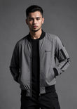 A man wearing a gray and black bomber jacket with a black crewneck shirt and black pants, standing against a dark gray studio background. The jacket features a small embroidered logo on the left chest that reads 'SOLOMON'.