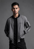 A man wearing a gray and black bomber jacket with a black crewneck shirt and black pants, standing against a dark gray studio background. The jacket features a small embroidered logo on the left chest that reads 'SOLOMON'.