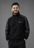 A man wearing a black zip-up track jacket with dark gray sleeve panels and a small white 'Soliches' logo on the left chest, standing against a solid gray studio background.