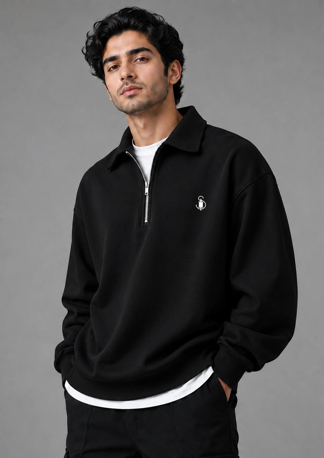 A man wearing a black half-zip sweatshirt with a small white embroidered logo on the left chest, layered over a white t-shirt and black pants, photographed against a solid gray studio background.