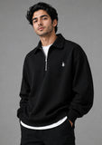 A man wearing a black half-zip sweatshirt with a small white embroidered logo on the left chest, layered over a white t-shirt and black pants, photographed against a solid gray studio background.